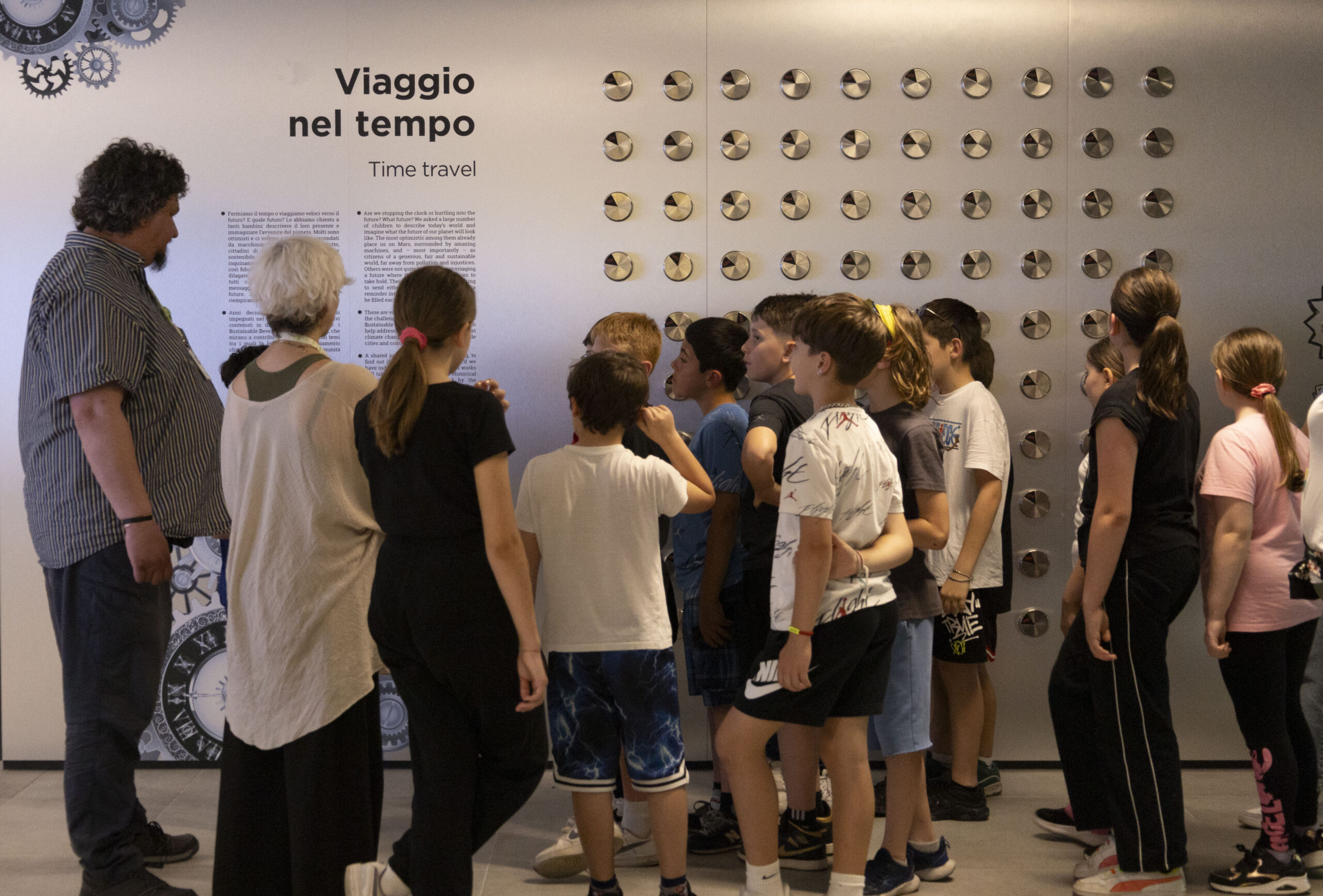 Un gruppo di bambine e bambini della scuola primaria ascolta un’operatrice che racconta il progetto “Capsule del Tempo”, indicando la parete espositiva con le capsule d’acciaio custodite a Heritage Lab.
