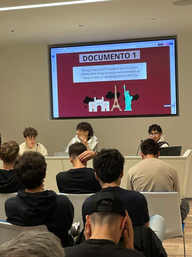 Studenti durante una presentazione all'Heritage Lab. Tre ragazzi siedono al tavolo dei relatori, mentre sullo schermo appare una slide intitolata “DOCUMENTO 1” con il tema “Smog e monumenti”. La slide propone un’attività divulgativa sull’effetto dello smog sui monumenti, illustrata con immagini della Torre Eiffel, dell’Arco della Pace e della Statua della Libertà. In primo piano, altri studenti ascoltano la presentazione.